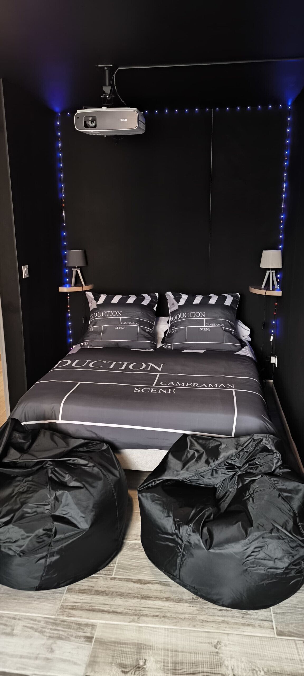 Une chambre à coucher avec un lit recouvert d’une couette rayée noire et blanche et orné d’oreillers sur lesquels sont inscrits les mots « PRODUCTION », « CAMÉRAMAN » et « SCÈNE ». On y trouve deux fauteuils en forme de haricot noirs posés sur le sol, un projecteur fixé au mur et deux petites lampes de chaque côté du lit.
