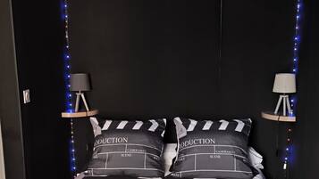 Une chambre à coucher avec un lit recouvert d’une couette rayée noire et blanche et orné d’oreillers sur lesquels sont inscrits les mots « PRODUCTION », « CAMÉRAMAN » et « SCÈNE ». On y trouve deux fauteuils en forme de haricot noirs posés sur le sol, un projecteur fixé au mur et deux petites lampes de chaque côté du lit.