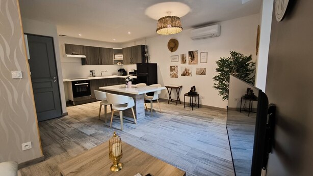 Une cuisine moderne avec des meubles de couleur sombre, un réfrigérateur noir et un plan de travail blanc. On y trouve également un coin repas avec une table en bois et des chaises blanches.