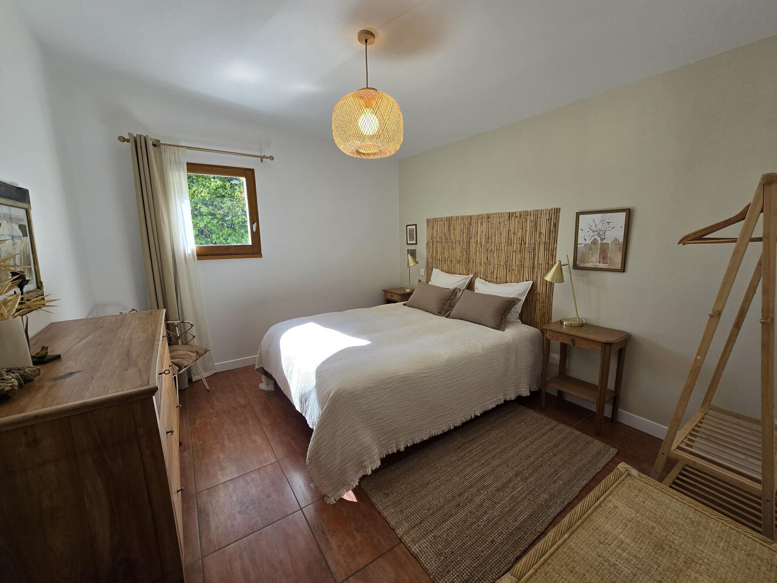 A bedroom with a bed, bedside tables, a hanging light fixture, a window with curtains, and a wooden floor.