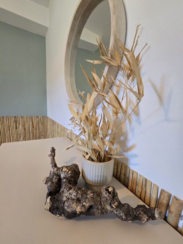 Una habitación con una mesa blanca, una planta decorativa en una maceta blanca, un espejo en la pared y un trozo de madera a la deriva.