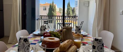 Una mesa en el balcón con alimentos para el desayuno, un jarrón y vistas a edificios y árboles.