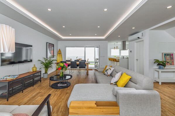 A modern living room with a grey sofa, a coffee table, and a TV. There is a dining area with chairs and a table, and a kitchen with a white cabinet and a plant.