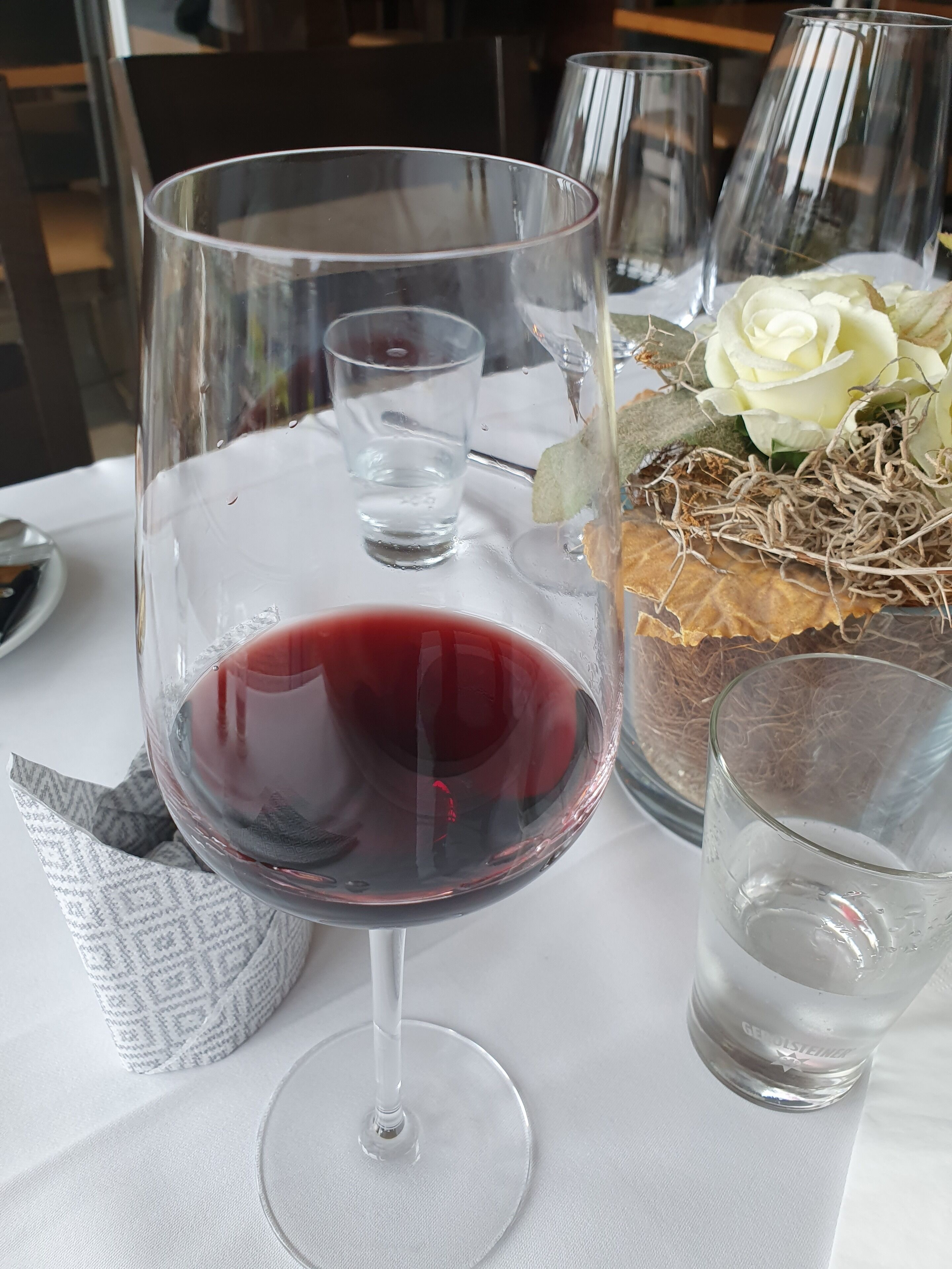 A table setting with a wine glass filled with red wine, a clear glass, and a decorative centerpiece with a white rose and dried foliage.