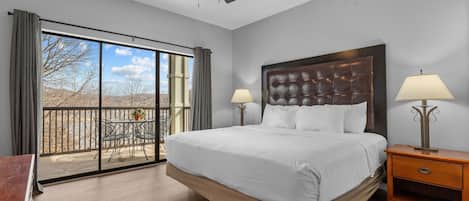 A spacious bedroom with a large bed, two bedside lamps, a ceiling fan, and a balcony with a table and chairs.
