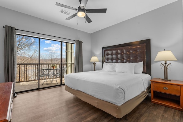 A spacious bedroom with a large bed, two bedside lamps, a ceiling fan, and a balcony with a table and chairs.