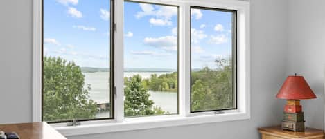 A bedroom with a large window offering a view of a lake and trees. There is a bedside table with a lamp and a wooden nightstand.