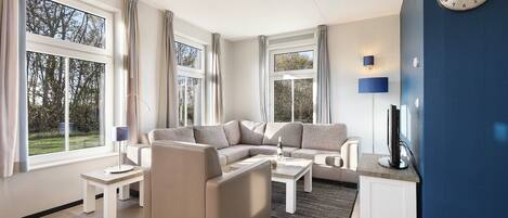 A modern living room with a sectional sofa, a coffee table, and a TV. There are large windows with curtains, a lamp, and a clock on the wall.
