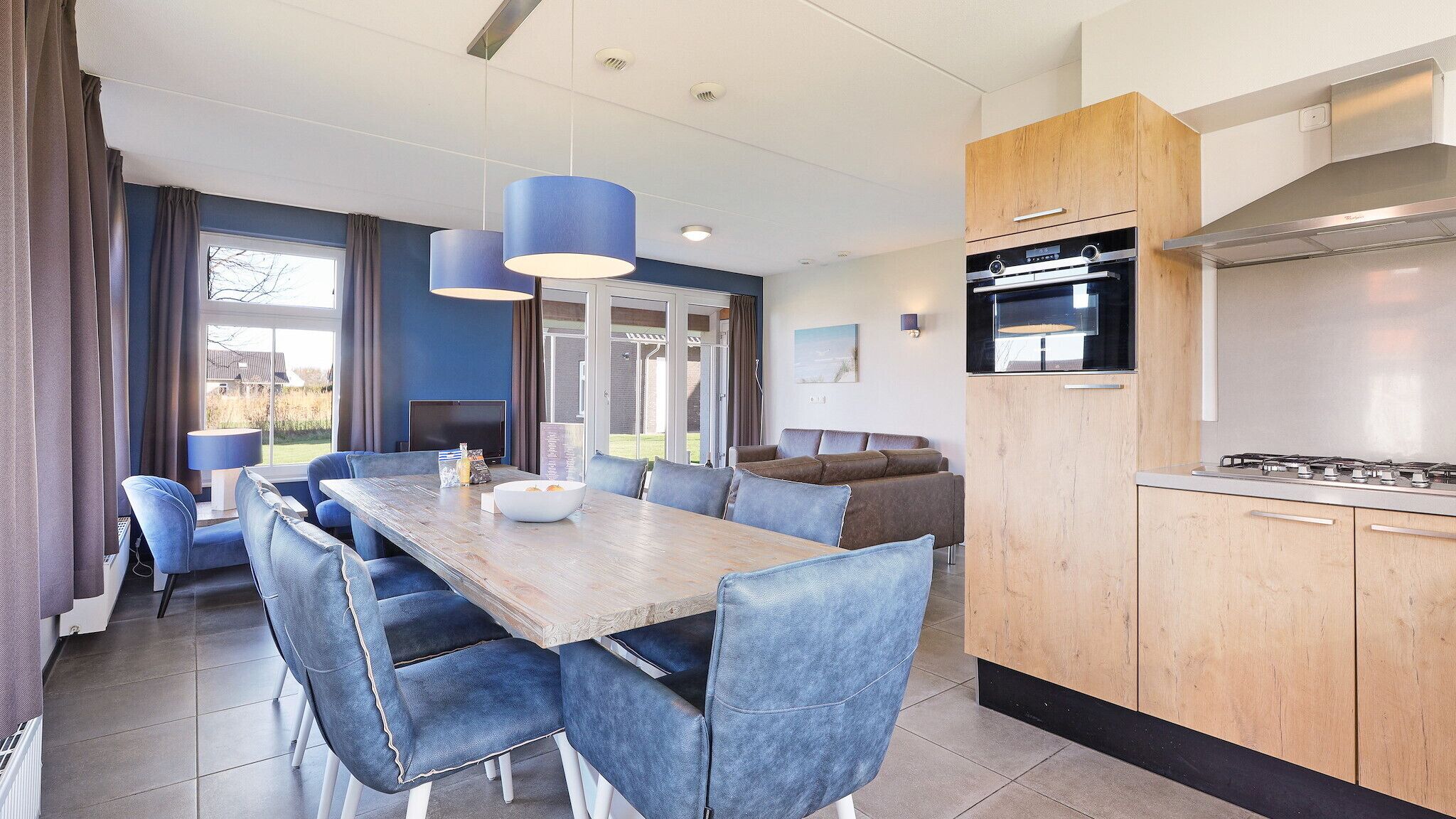 Une cuisine moderne dotée d’un four et d’une cuisinière encastrés, un coin repas avec des chaises bleues et une table en bois, et une grande fenêtre avec des rideaux.