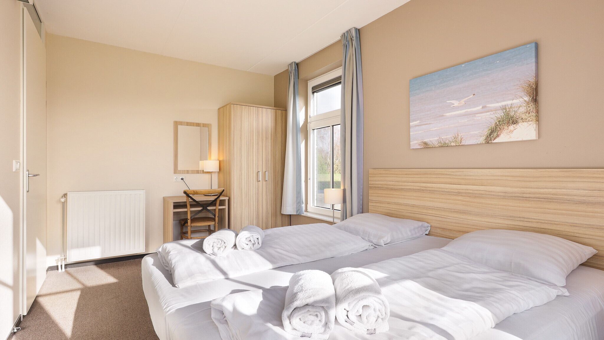 A hotel room with a bed, a desk, a chair, a radiator, a window with curtains, and a painting on the wall.