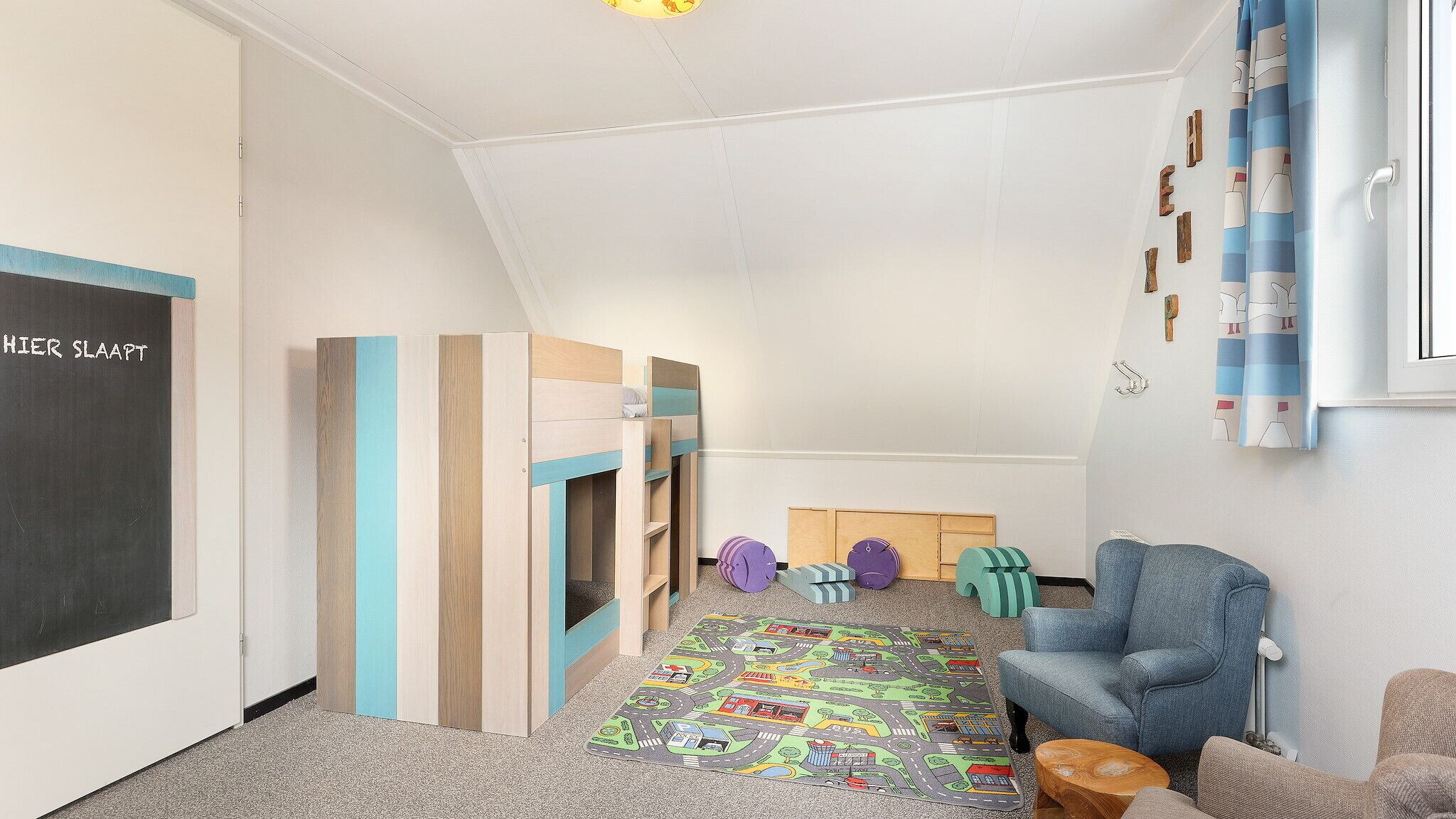 Een kindvriendelijke kamer met een schoolbord, houten speeltoestellen, een kleurrijke speelmat en een blauwe fauteuil.