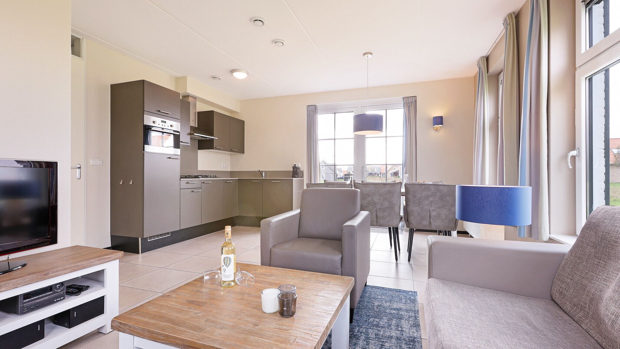 Una sala moderna con un televisor de pantalla plana, una mesa de centro de madera y un sofá. Hay una zona de cocina con armarios y un horno empotrado. La habitación tiene ventanas grandes con cortinas y vistas al exterior.