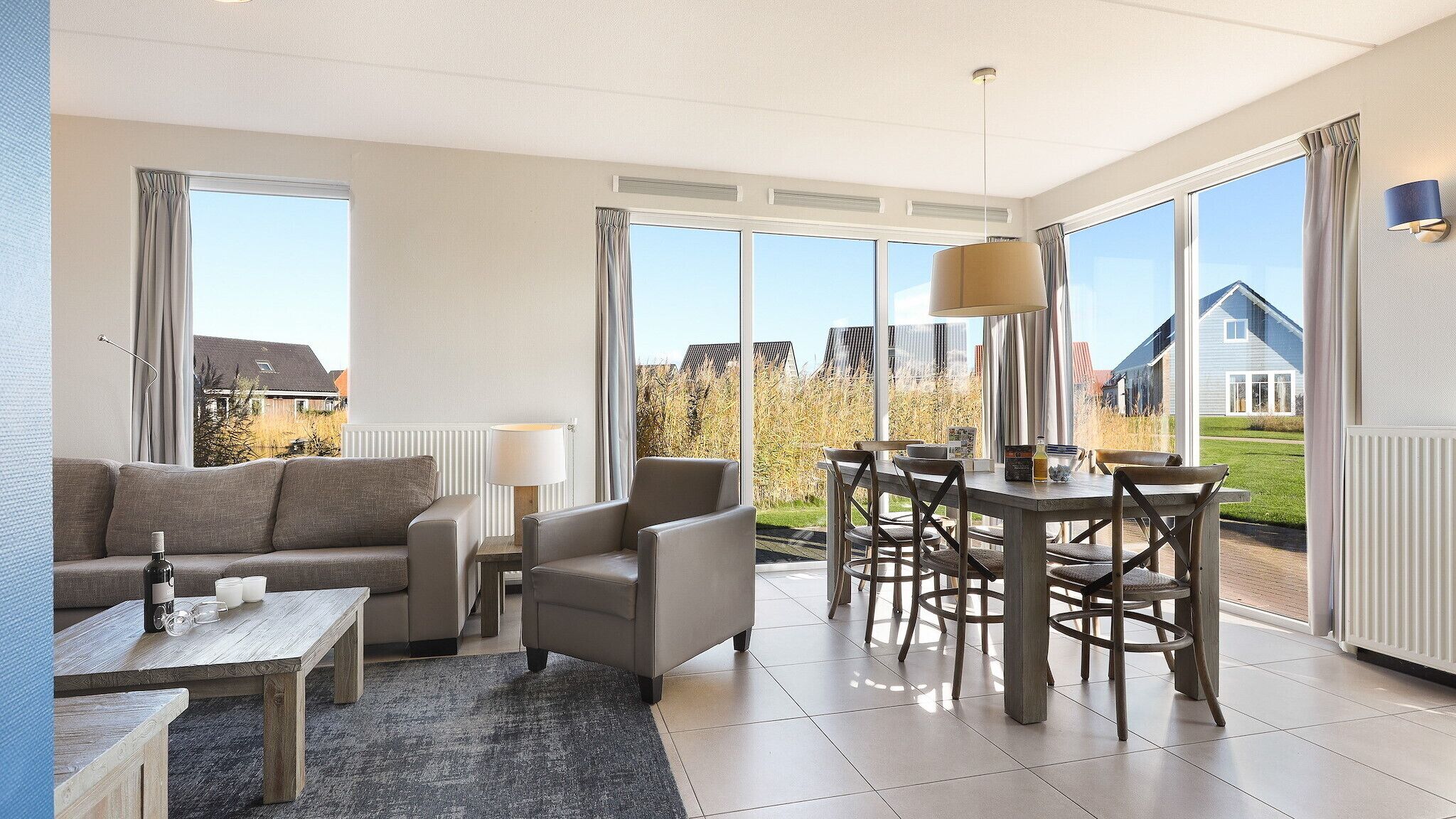 Ein modernes Wohnzimmer mit einer Couch, Sesseln, einem Couchtisch und einem Essbereich mit Tisch und Stühlen. Große Fenster bieten einen Ausblick nach draußen.
