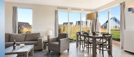 Ein modernes Wohnzimmer mit einer Couch, Sesseln, einem Couchtisch und einem Essbereich mit Tisch und Stühlen. Große Fenster bieten einen Ausblick nach draußen.