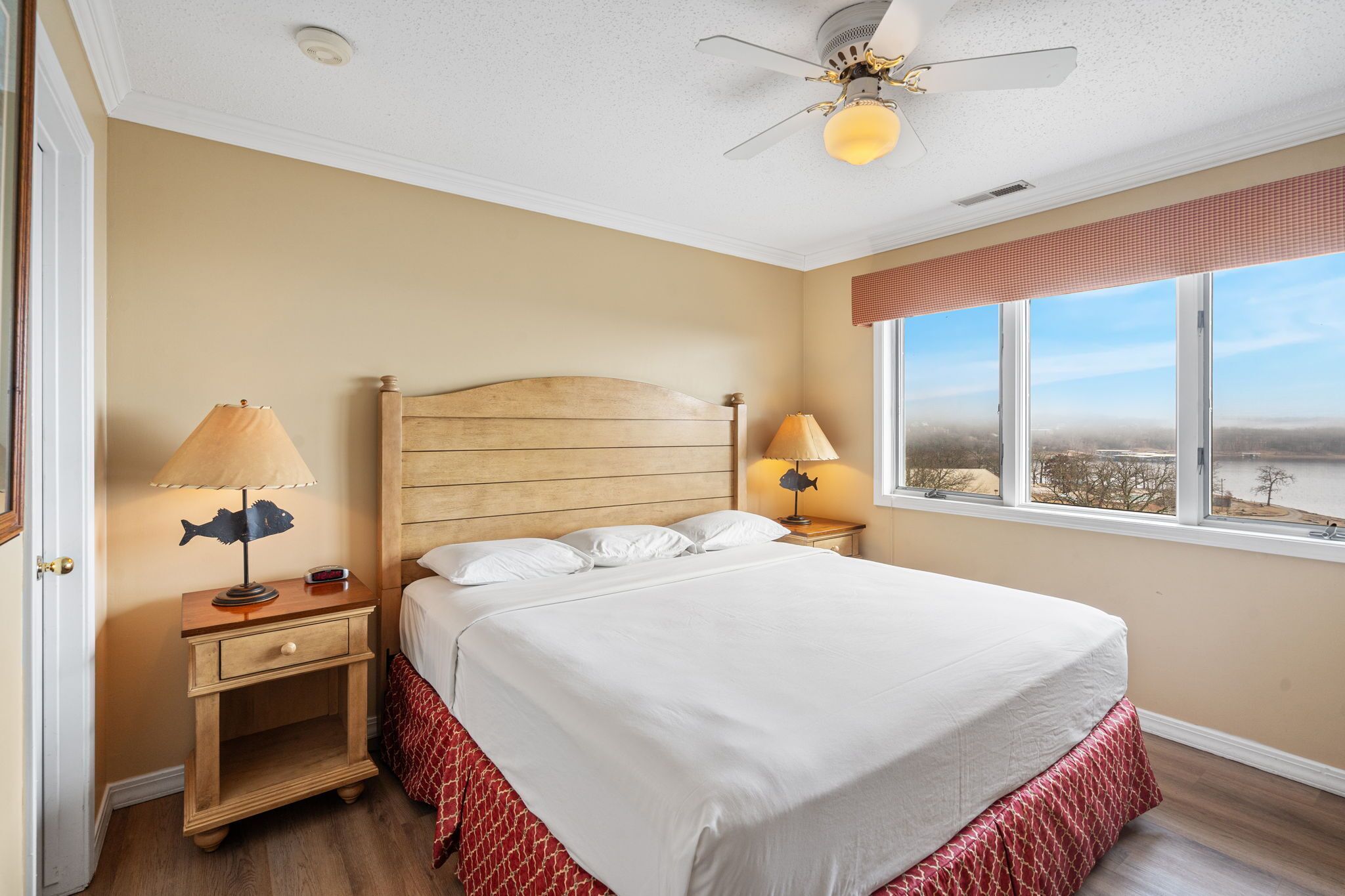 A bedroom with a wooden headboard, two bedside lamps, a view of a body of water through a window, and a ceiling fan.
