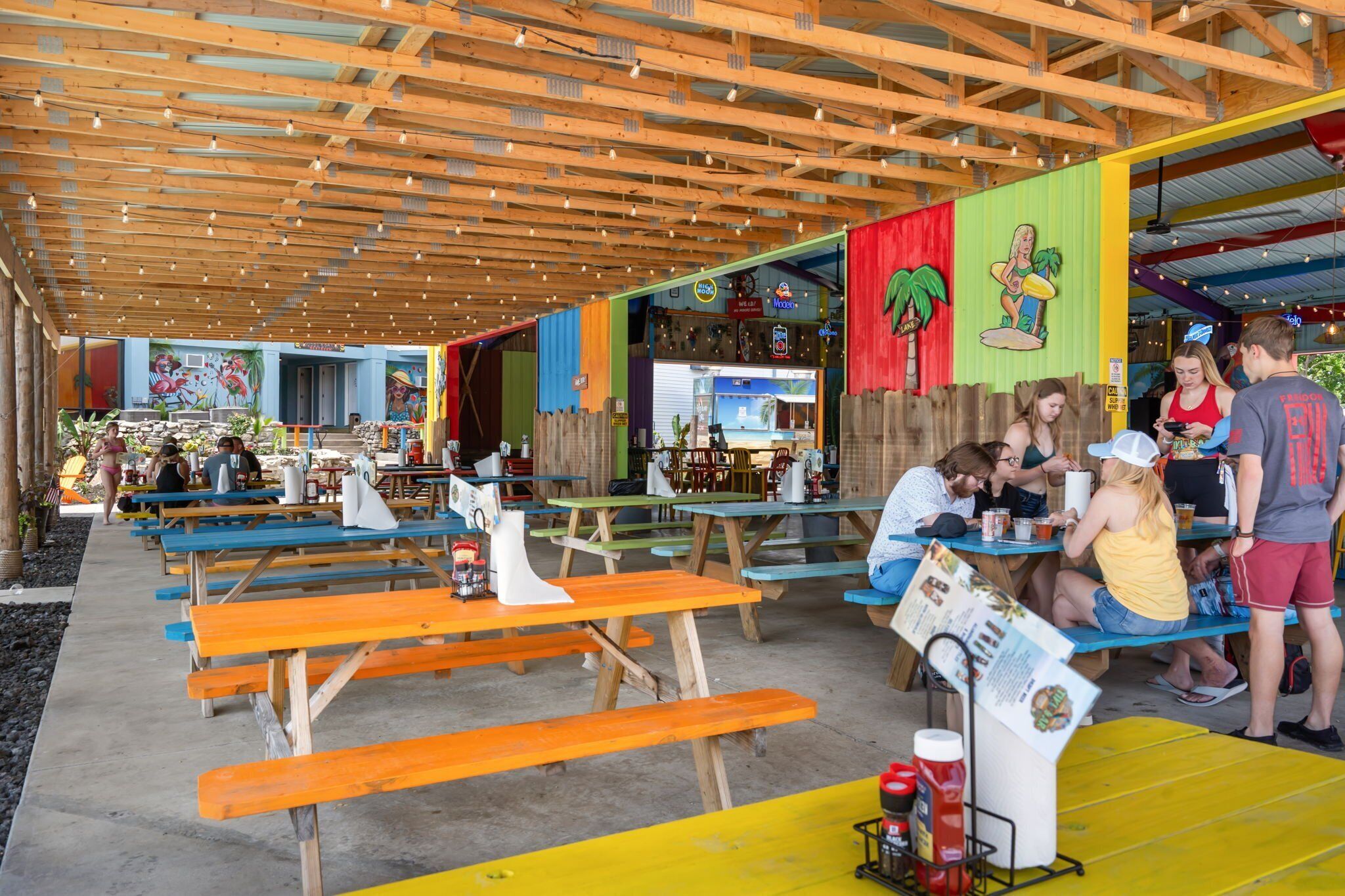 An outdoor dining area with wooden tables and benches, a covered seating area, and a colorful mural on the wall.