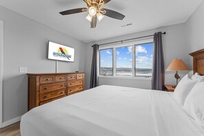 Une chambre à coucher comprenant un lit, une commode en bois, un ventilateur de plafond, une télévision, une fenêtre avec des rideaux et une lampe.