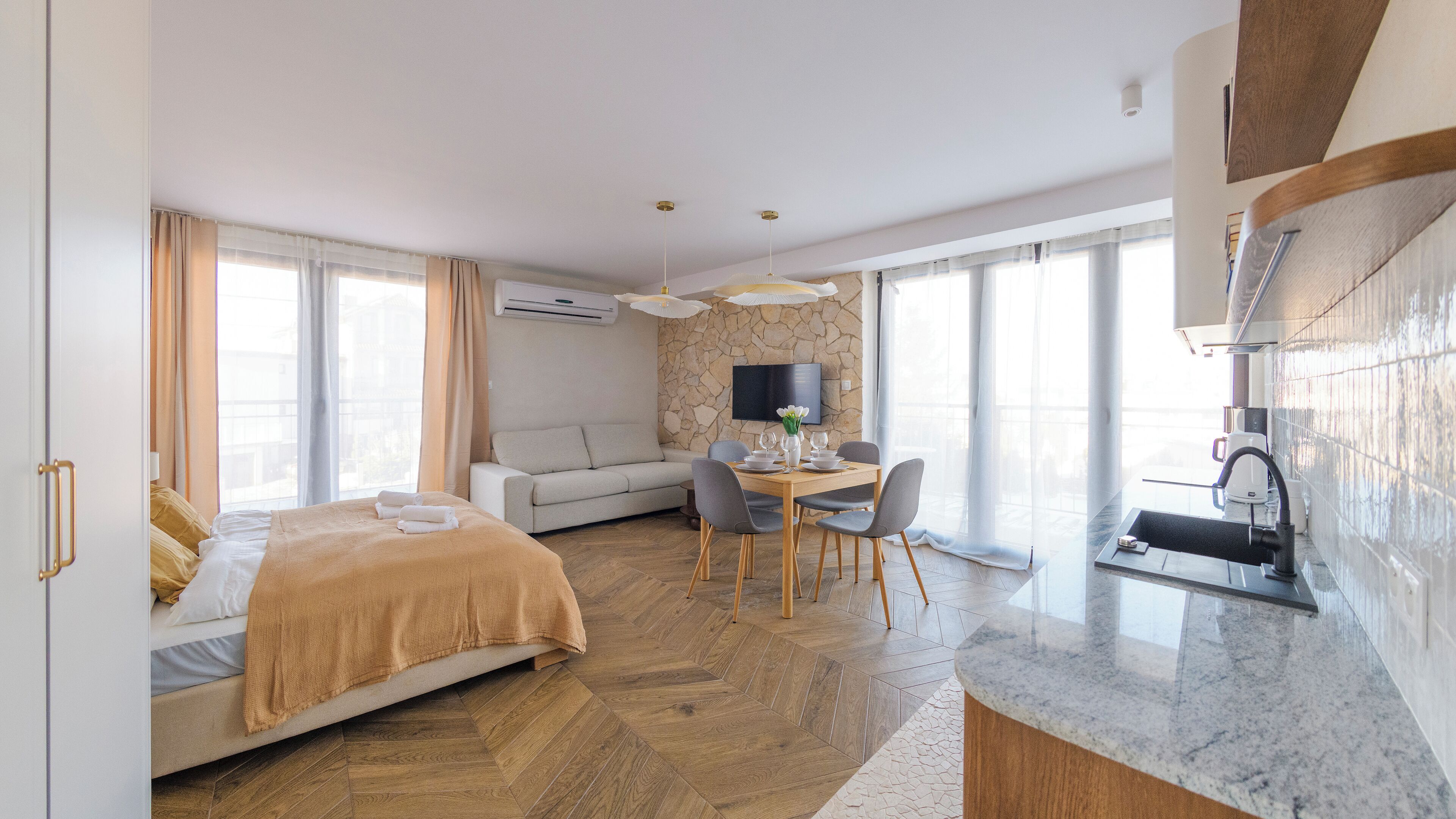 A modern hotel room with a bed, a sofa, a dining table with chairs, a TV, and a kitchen area.