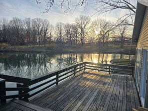 A wooden deck with a railing overlooking a calm body of water, surrounded by trees and a building. - Charming Cabin on Private Pond – 2 Miles from Lake Rathbun, Southern Iowa (Centerville)