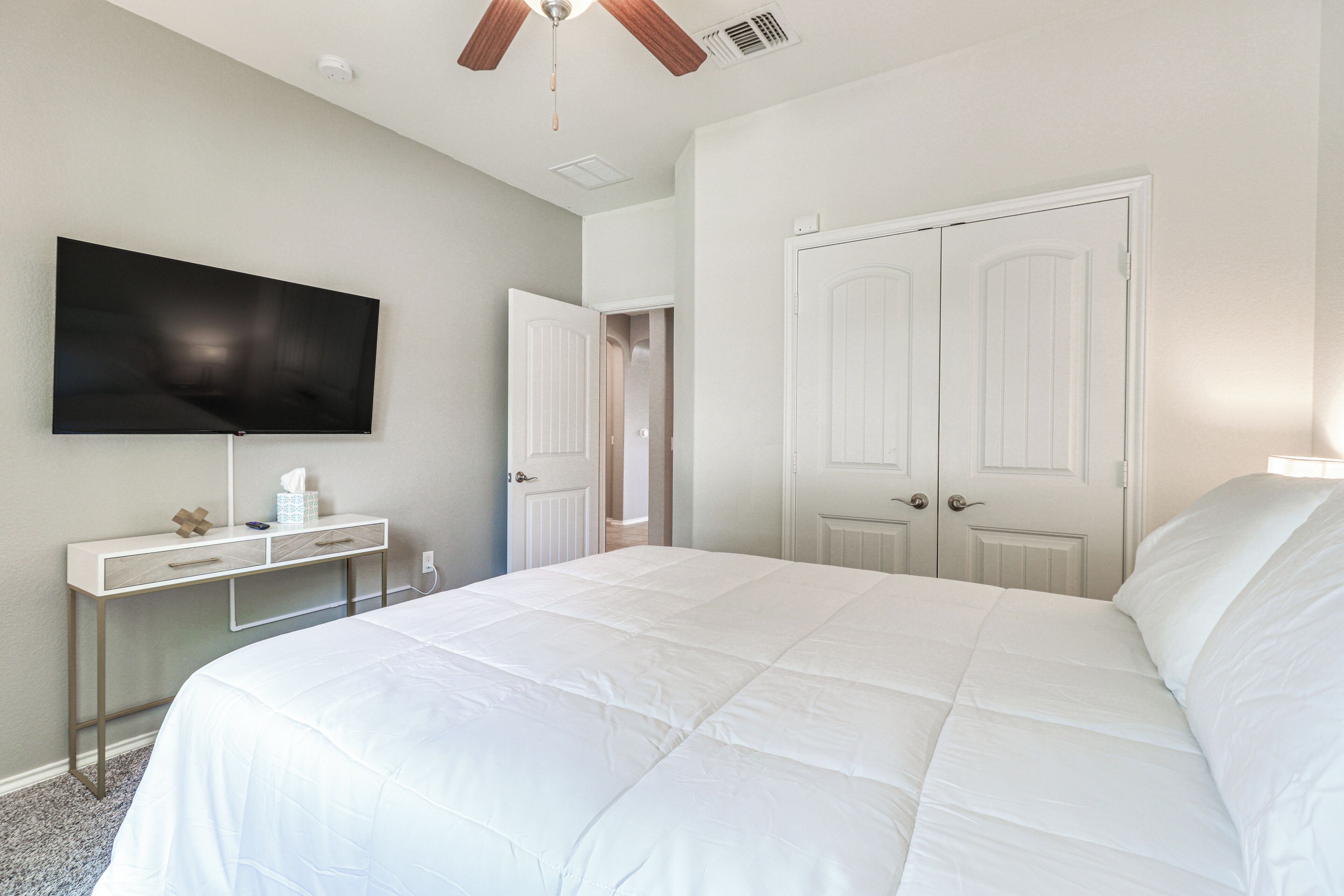 A bedroom with a large bed, a TV mounted on the wall, a desk with drawers, and a wardrobe with two doors.