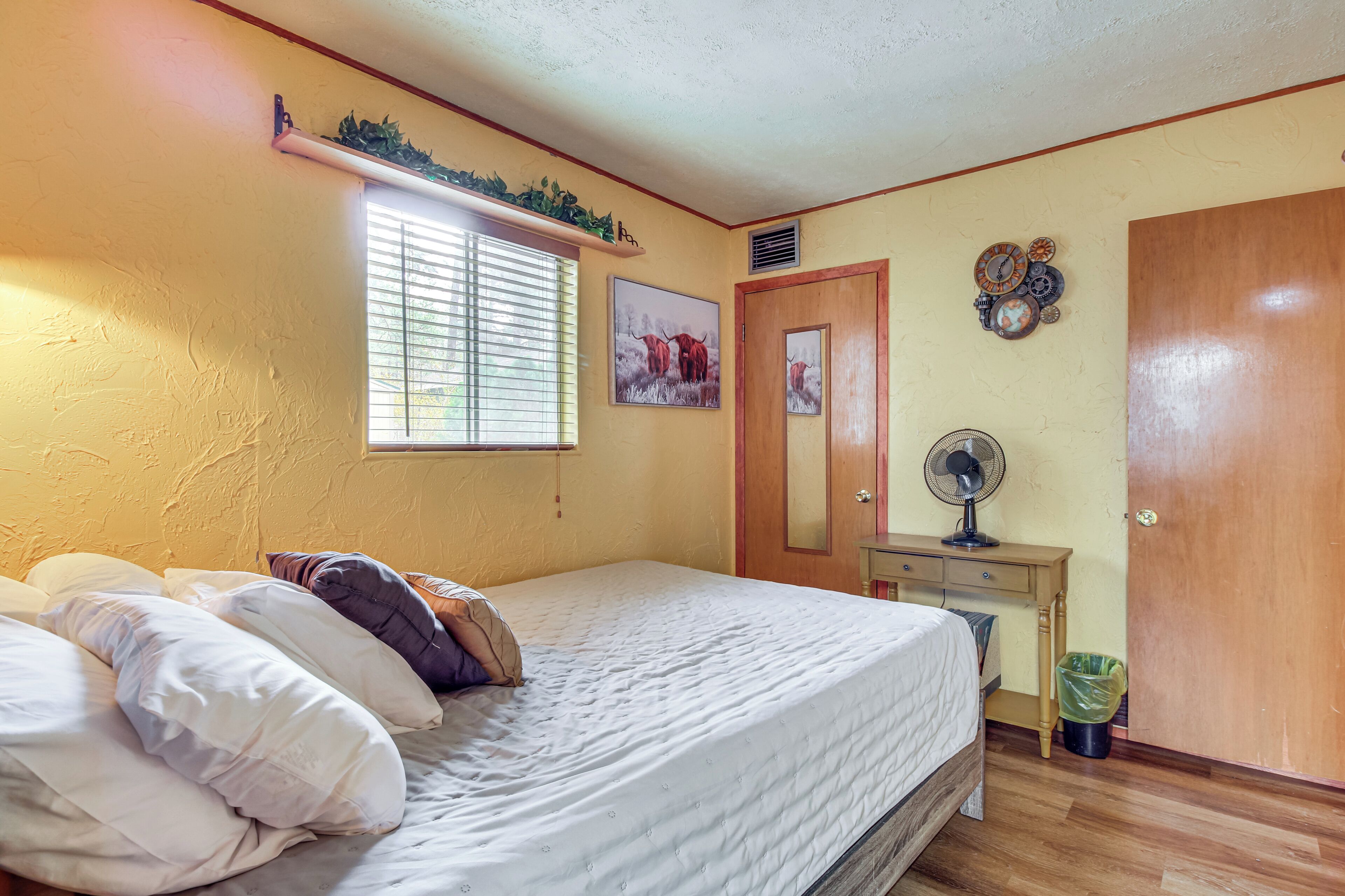 A bedroom with a bed, a window with blinds, a wall clock, a small table with a fan, and a door.