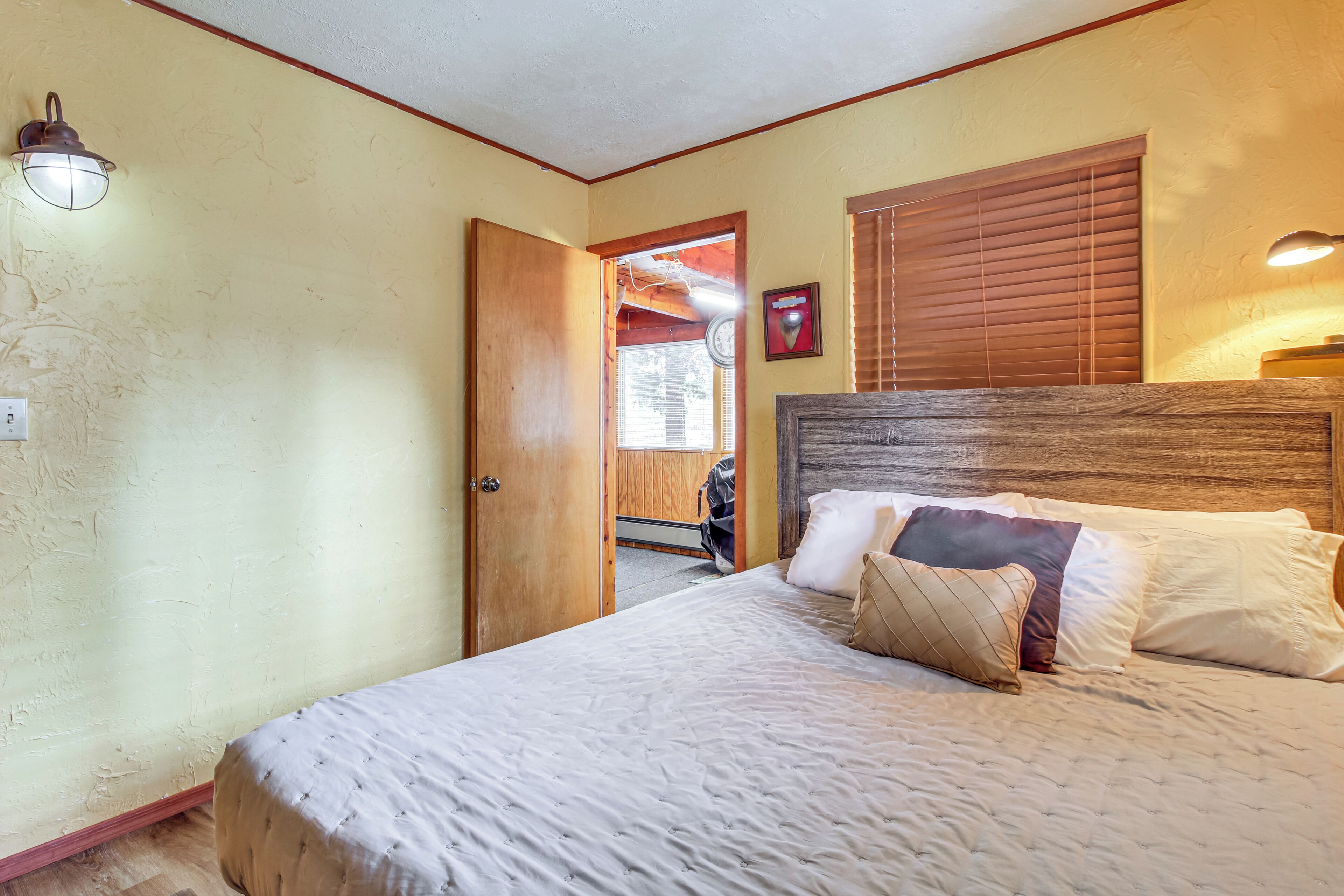 Un dormitorio con cama, cabecera de madera, mesita de noche, reloj de pared y puerta que da a otra habitación.