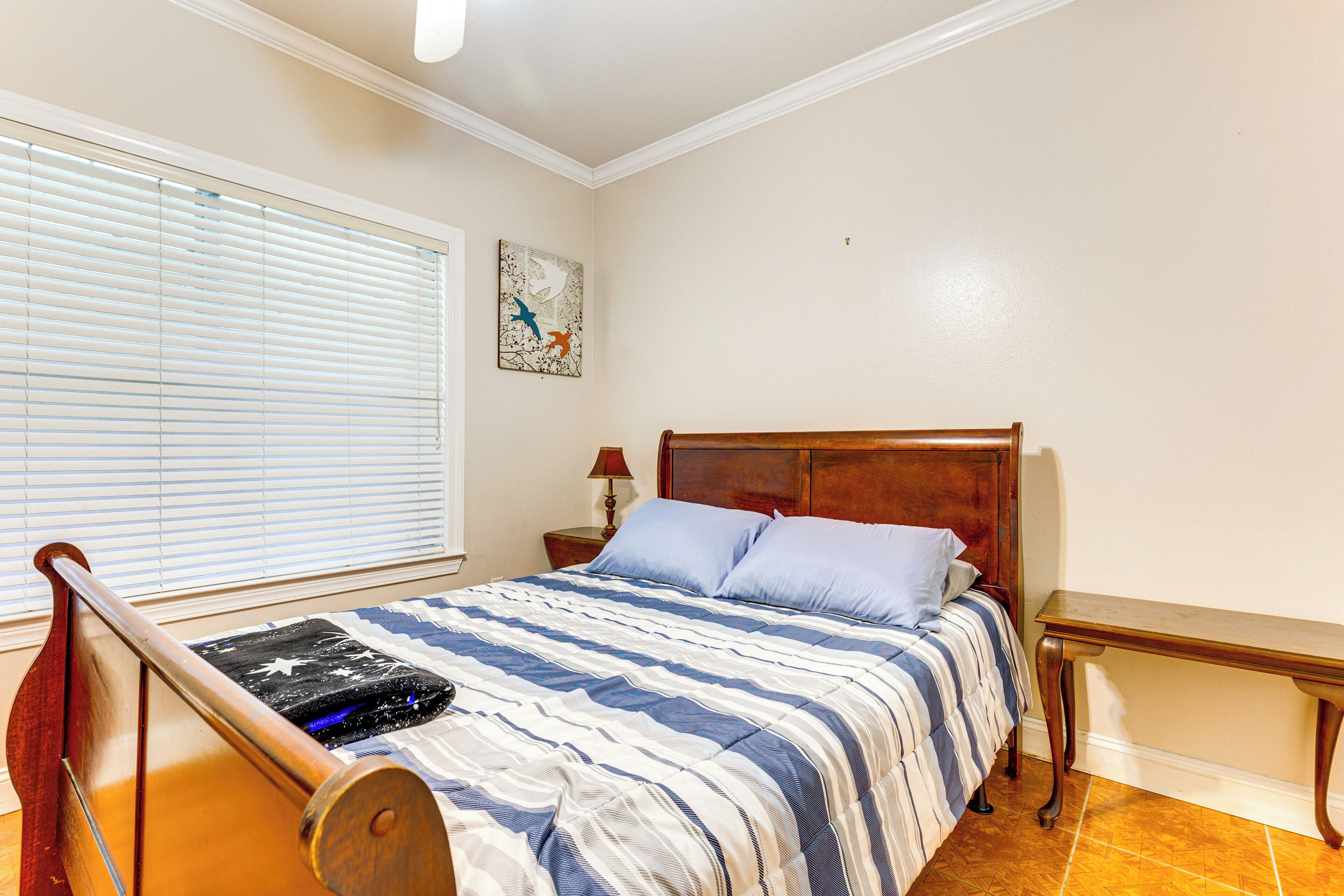 A bedroom with a bed, a nightstand, a wooden bench, a window with blinds, and a wall-mounted artwork.