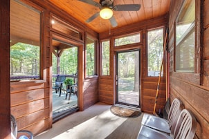 A cabin interior with wooden walls, large windows, a ceiling fan, and a porch area with outdoor seating.