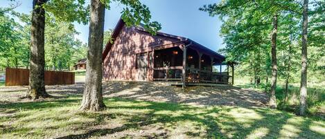 A rustic cabin with a porch, surrounded by trees and a gravel path.