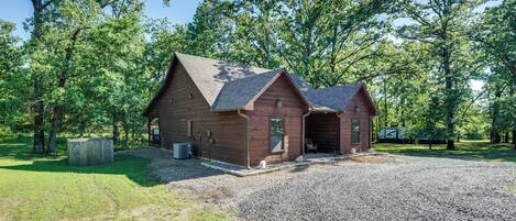 A rustic cabin with a gravel driveway and a covered outdoor seating area, surrounded by trees.