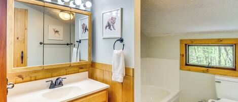 A bathroom with a wooden vanity, a large mirror with lights, a bathtub, and a window.