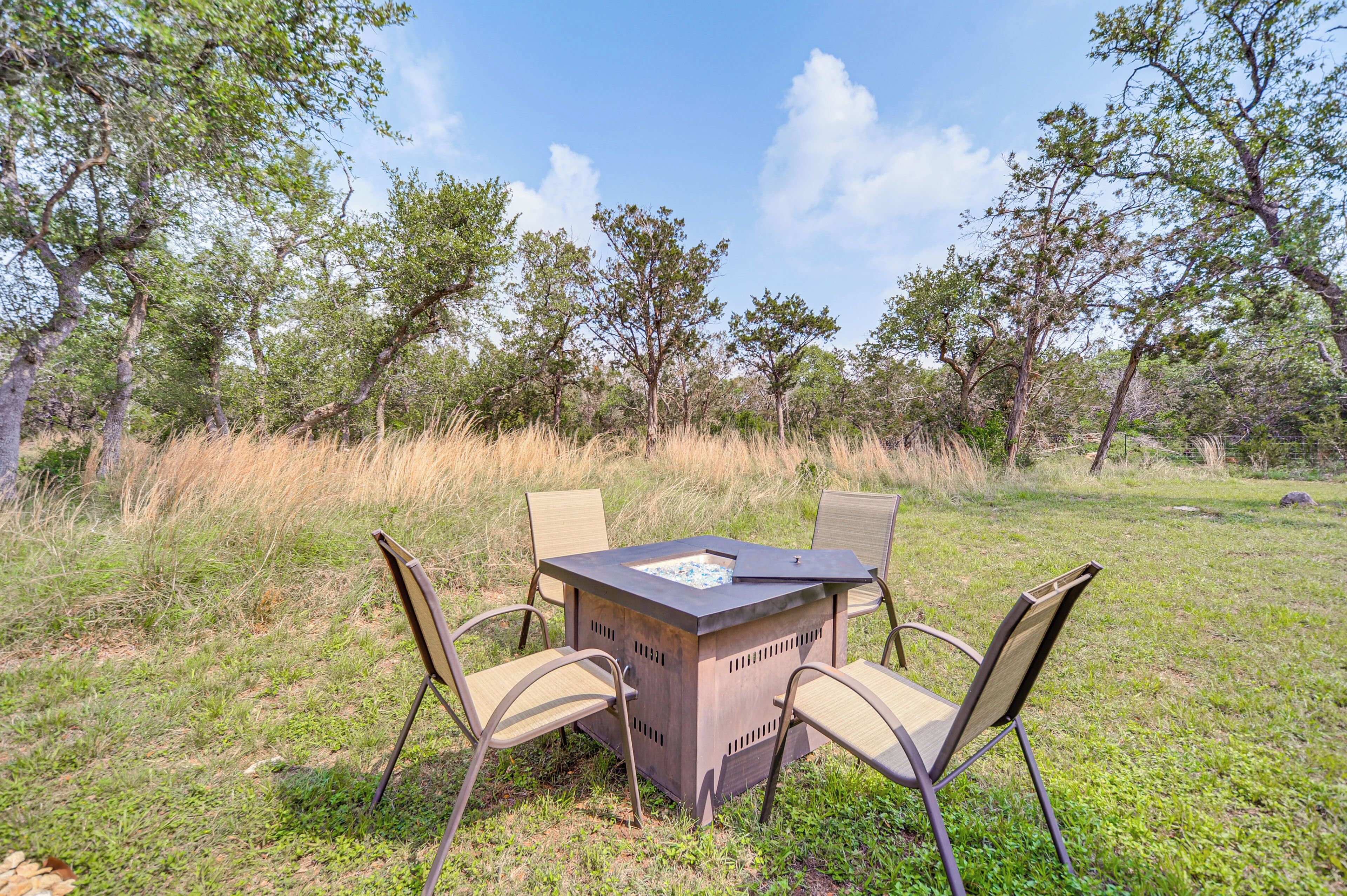 An outdoor seating area with a square table and four chairs, surrounded by grass and trees.