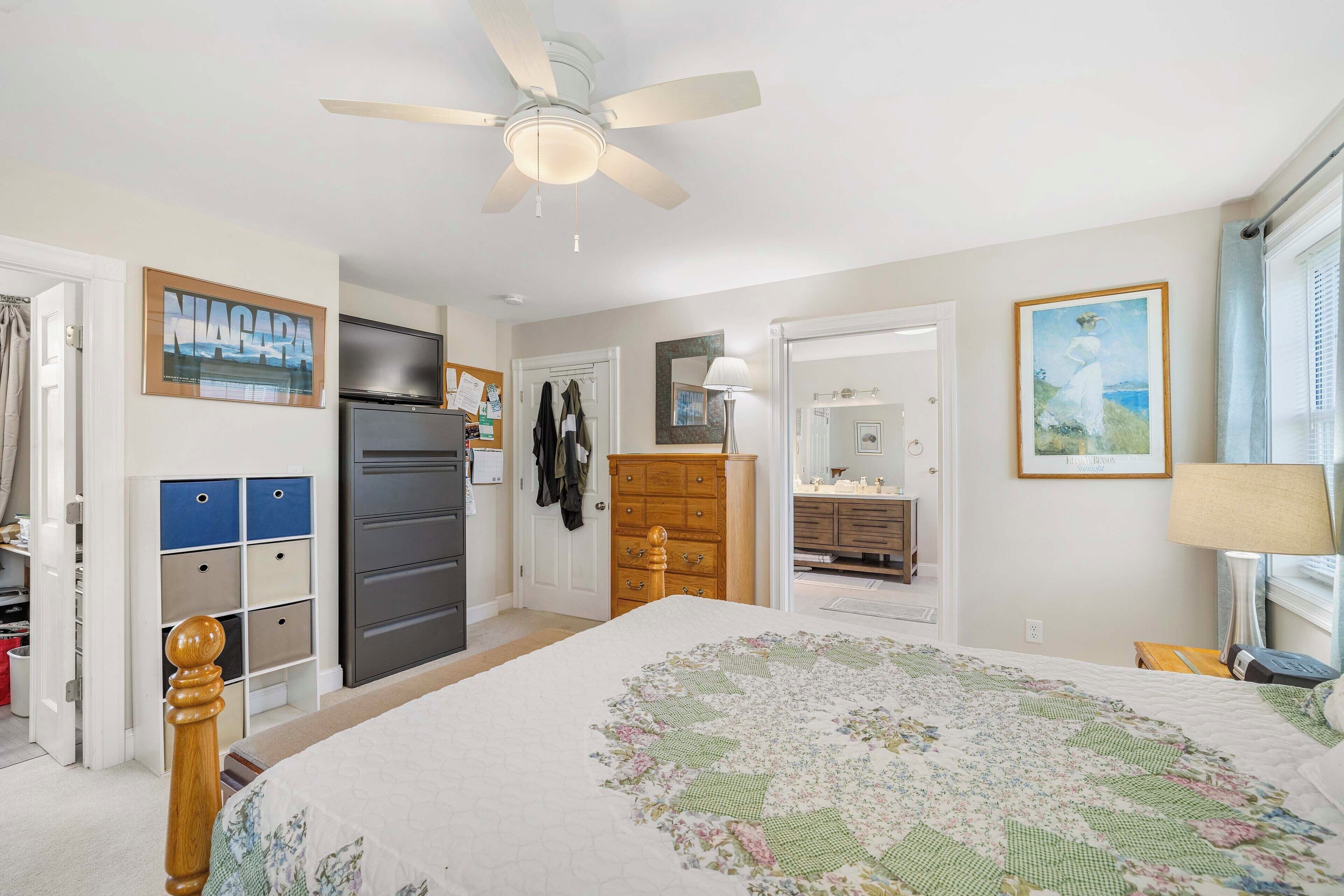 A bedroom with a bed, a ceiling fan, a dresser, a wardrobe, and a door leading to another room.