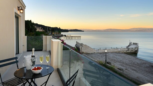A balcony with a table set for two, a bottle of champagne, and two glasses of wine. The balcony overlooks a calm sea and a pier extending into the water. - Beachfront Achillion Bay Villa in Corfu (Corfu)