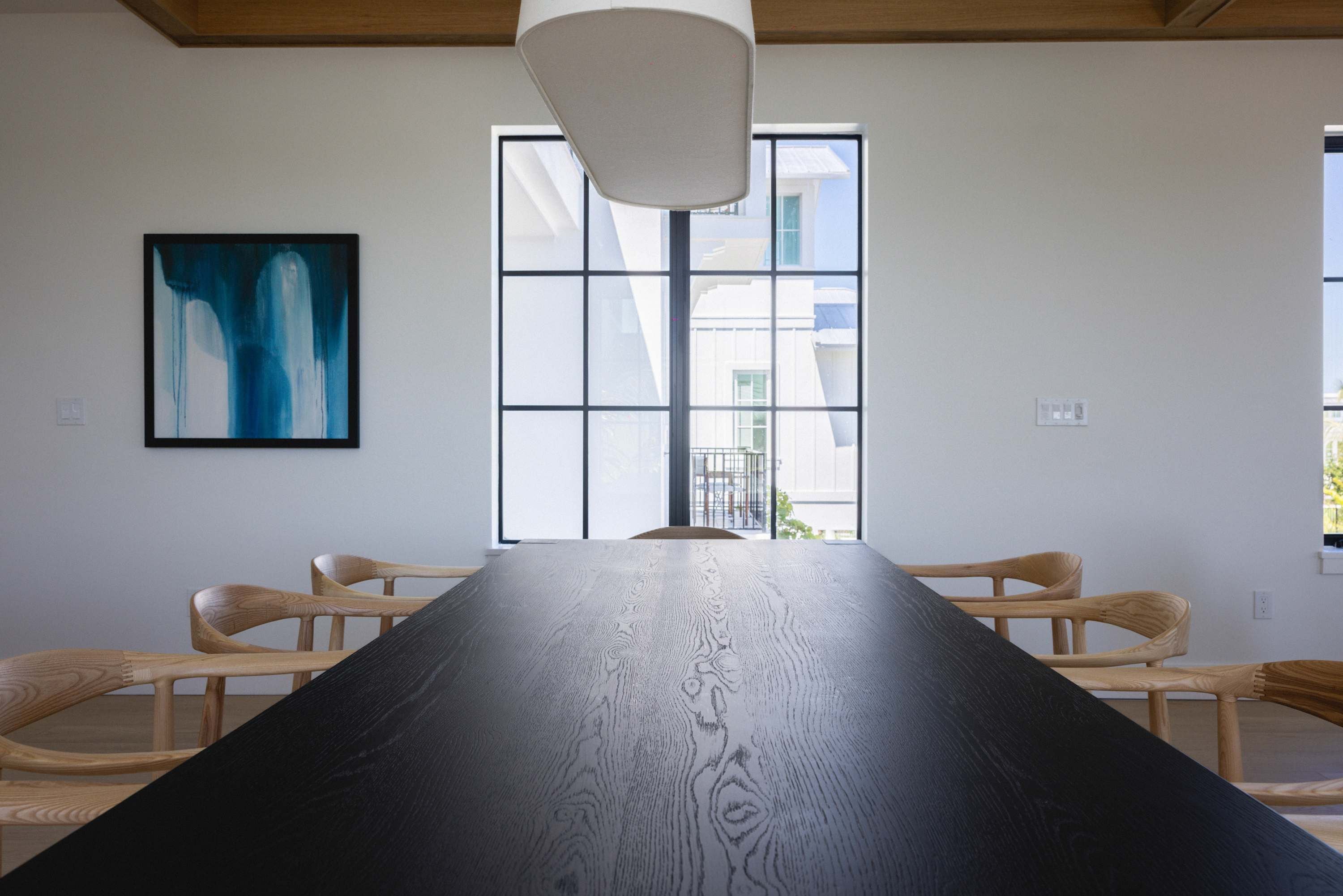 Un salón de conferencias con una mesa larga, dos sillas de madera, una lámpara colgante moderna y un ventanal con vistas a edificios.