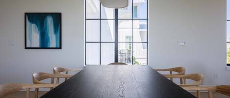 Un salón de conferencias con una mesa larga, dos sillas de madera, una lámpara colgante moderna y un ventanal con vistas a edificios.