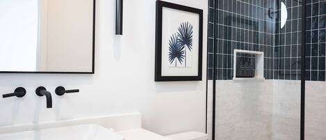 Un baño moderno con un lavamanos blanco, grifos negros, una obra de arte con una planta enmarcada y una zona de ducha con azulejos negros.