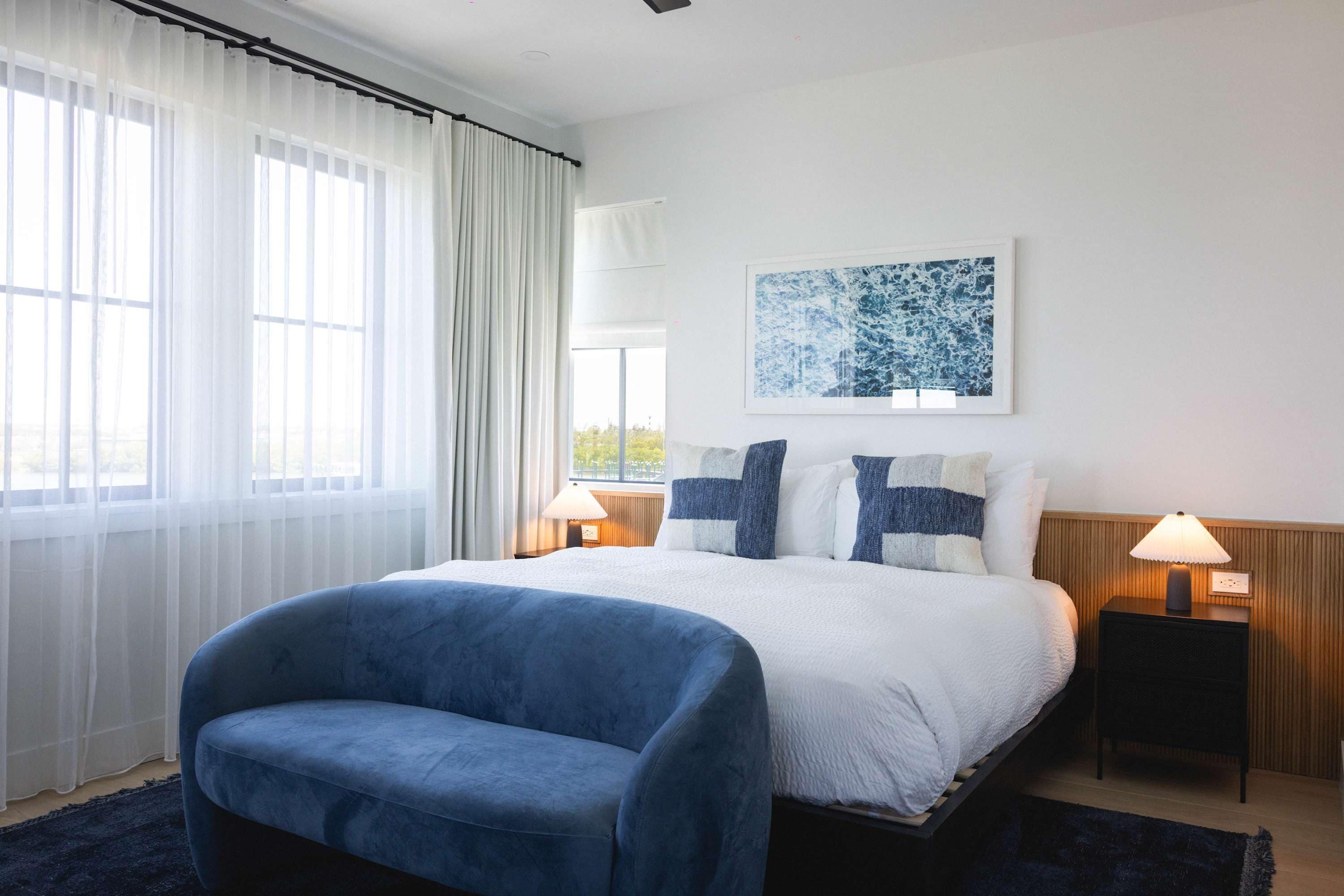 A hotel room with a bed, a blue armchair, a window with sheer curtains, a wall-mounted artwork, and two bedside lamps.