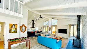 A spacious living room with a blue sofa, a foosball table, a fireplace, and a dining area with a kitchen in the background. - Modern Yosemite Cabin, 15min Bass Lake, Sleeps 8 (Oakhurst)