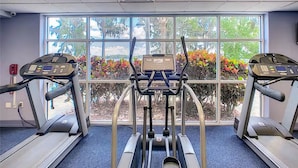 Ein Fitnessstudio mit drei Laufbändern, einem zentralen Crosstrainer und einem großen Fenster mit Blick auf Bäume und Pflanzen.