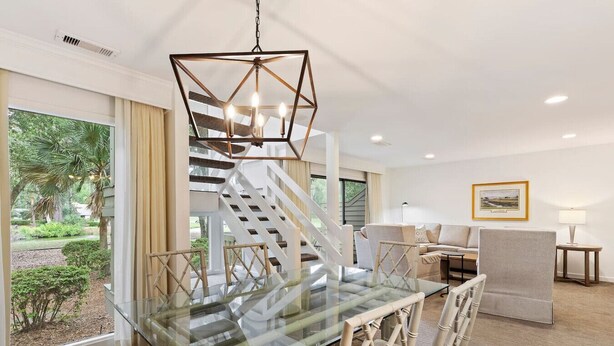 A modern dining area with a glass table and chairs, a geometric chandelier, a staircase, and a view of the outdoors. - 2879 - 2BR Villa | Golf Views | Sea Pines Pool (Hilton Head Island)