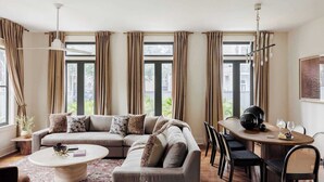 Un soggiorno moderno con un divano componibile, un tavolino rotondo e un tavolo da pranzo con sedie. Ampie finestre con tende offrono luce naturale e vista sull'esterno.