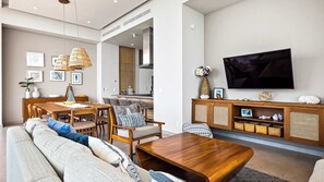 Ein modernes Wohnzimmer mit einer Couch, Couchtisch, Esstisch und Stühlen. Ein Flachbildfernseher ist an der Wand befestigt, und im Hintergrund ist eine Küche zu sehen.