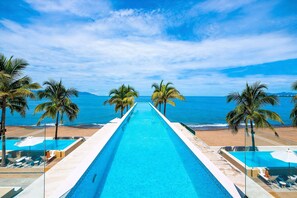 A long swimming pool with a beach view, surrounded by palm trees.