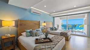 Ein Hotelzimmer mit einem großen Bett, einem Nachttisch, einer Lampe, einem Tablett mit einer Kerze und Blick auf den Strand und das Meer durch eine Schiebetür.