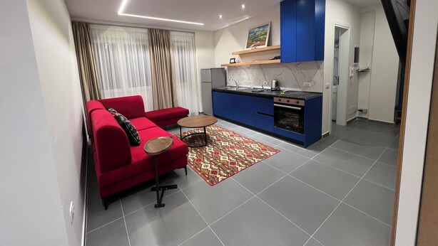 A modern living room with a red sofa, a small coffee table, and a patterned rug. Adjacent is a compact kitchen with blue cabinets and a built-in oven. - The Velvet Nest (Tirana)
