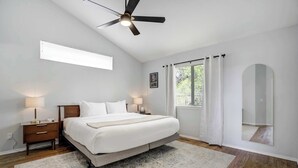 A bedroom with a bed, bedside tables, a ceiling fan, a window with curtains, and a mirror.