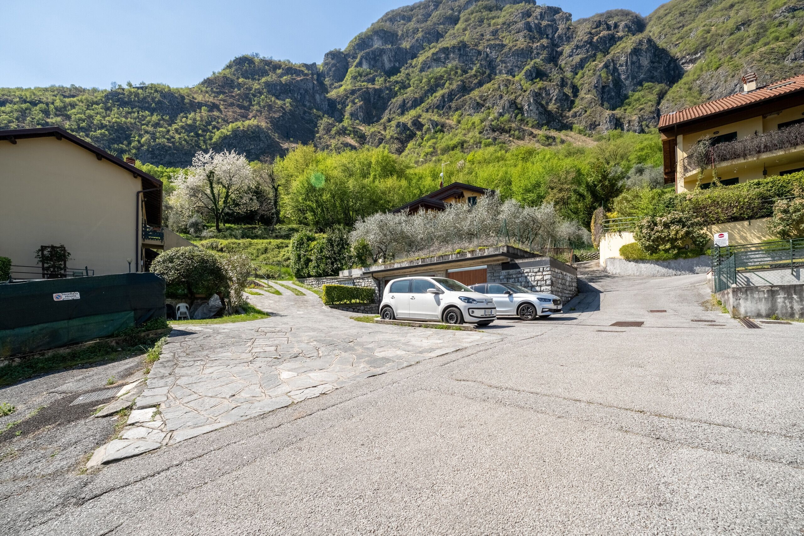 A residential street with a white car parked in the driveway, surrounded by lush greenery and mountains in the background.