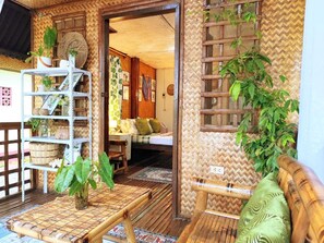 A room with a wooden floor, a bed with a green pillow, a shelf with plants, and a woven wall. - Amwani Garden Pool & Kubo Tent Rental (Puerto Princesa)