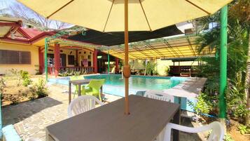 A pool area with a covered seating area, a table with chairs, and a building with a red roof in the background.